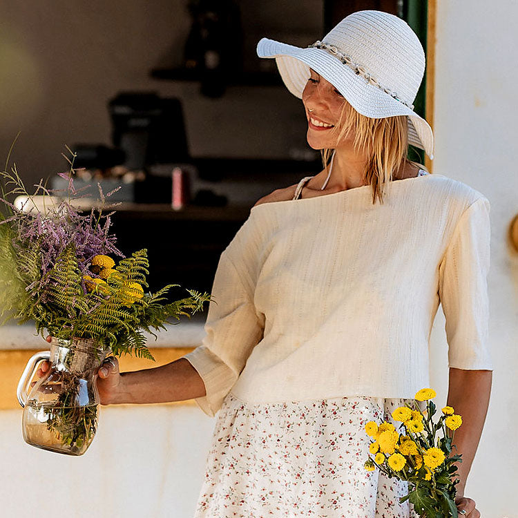 Frau mit weißem Sonnenhut hält bunte Blumensträuße vor rustikalem Haus in Portugal, sommerliche Hochzeitsinspiration