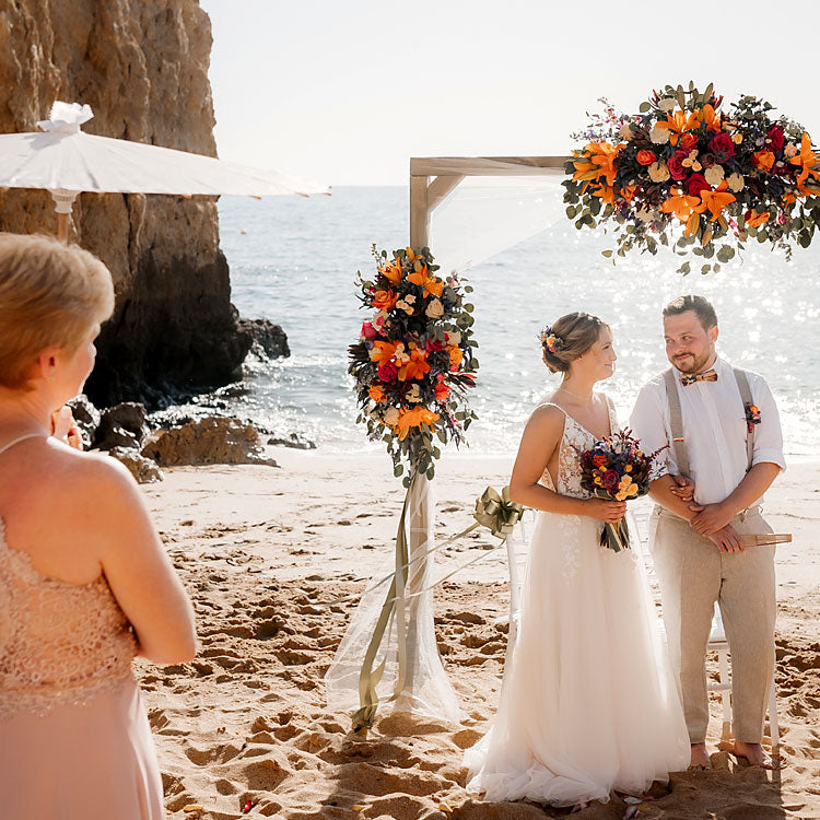 Brautpaar bei Strandhochzeit in Portugal, unter Blumenbogen mit Meerblick und bunter Blumendeko