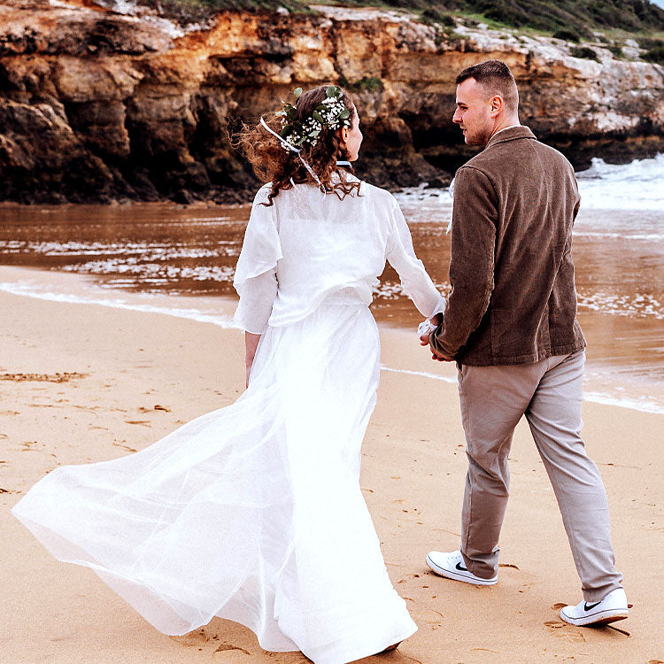 Brautpaar läuft Hand in Hand am Strand der Algarve entlang, Brautkleid weht im Wind vor Felsenkulisse