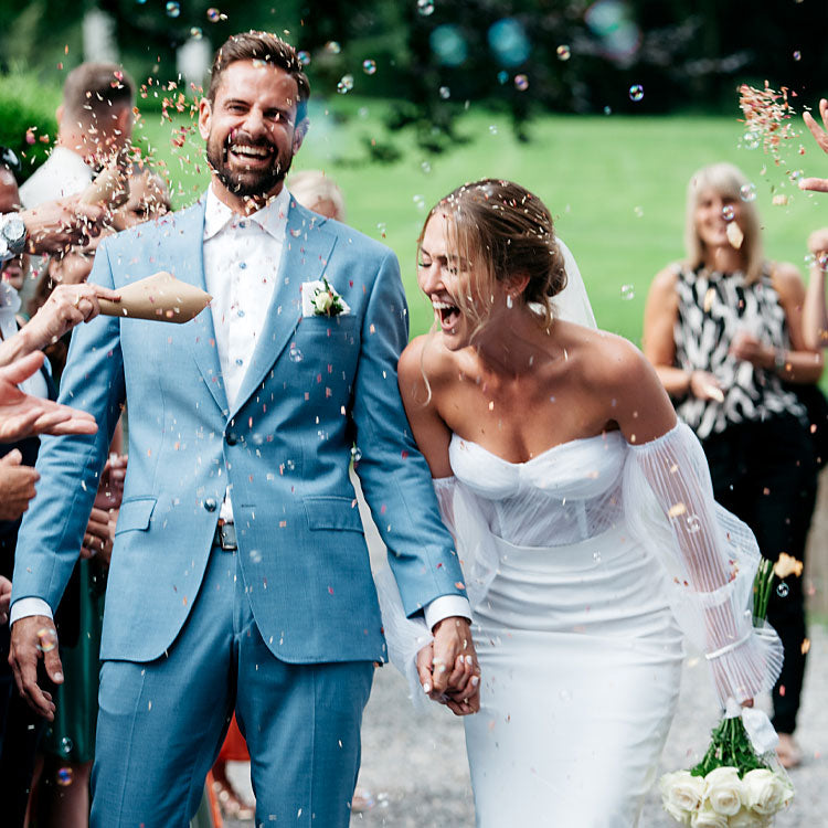 Brautpaar läuft lachend durch ein Spalier von Gästen, Konfetti und Seifenblasen bei der Hochzeitsfeier