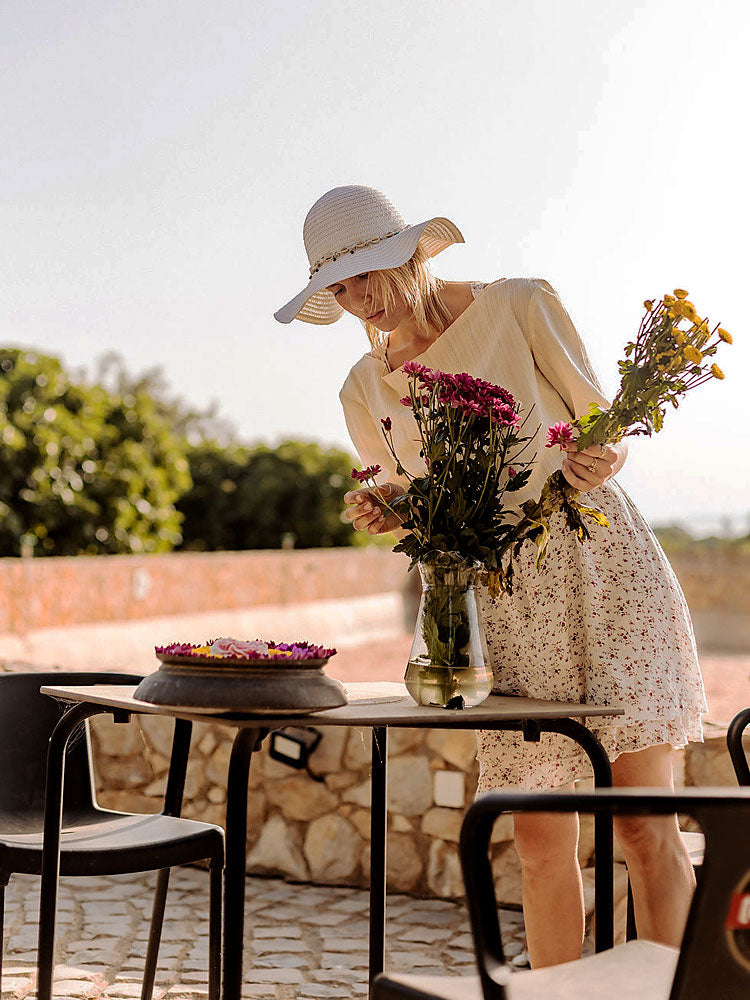 Frau mit weißem Sonnenhut arrangiert Blumen in einer Vase auf einem Tisch, sommerliche Hochzeitsdeko in Portugal