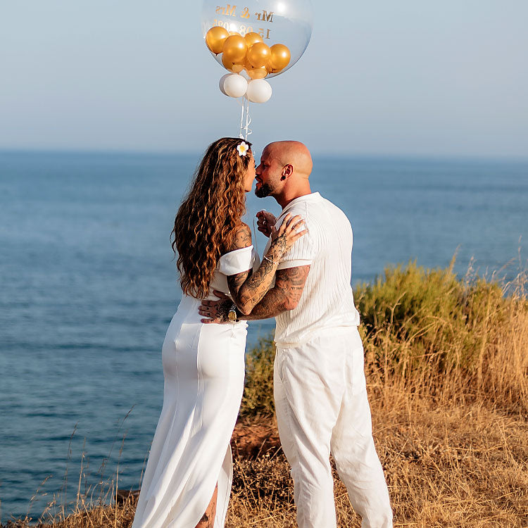 Brautpaar küsst sich bei freier Trauung an den Klippen der Algarve, mit goldenem Ballon und Meerblick