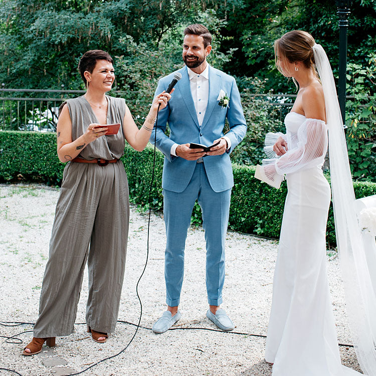 Freie Traurednerin hält Hochzeitsrede im Freien, Brautpaar steht lächelnd bei der Trauung im Garten