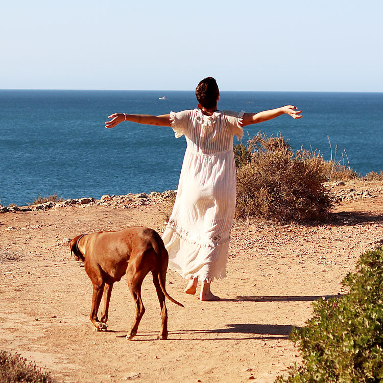 Susy im weißen Kleid läuft barfuß mit Hund an den Klippen der Algarve und blickt aufs Meer
